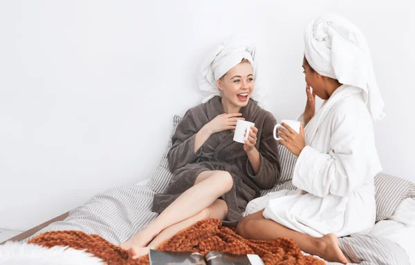 Joyful girlfriends in bathrobes sitting on bed and drinking tea — Stockfoto
