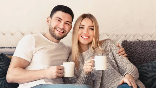 Jovem e sua esposa com duas xícaras de bebidas quentes relaxando em casa, panorama — Fotografia de Stock