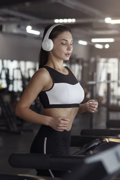 Pretty girl with headphones jogging on treadmill in gym — ストック写真