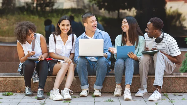 Radosni studenci uniwersytetu siedzący na kampusie z laptopem i śmiejący się razem — Zdjęcie stockowe