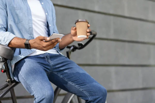 Nierozpoznawalny czarny facet siedzący na rowerze, relaksujący się przy smartfonie i kawie — Zdjęcie stockowe