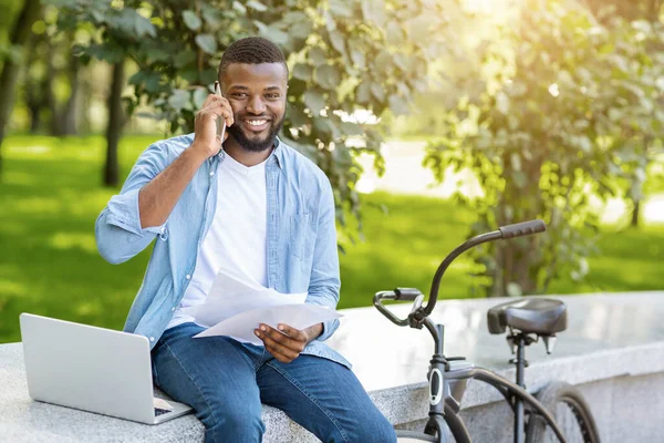 Sorrindo preto empresário com papéis e laptop conversando no celular ao ar livre — Fotografia de Stock