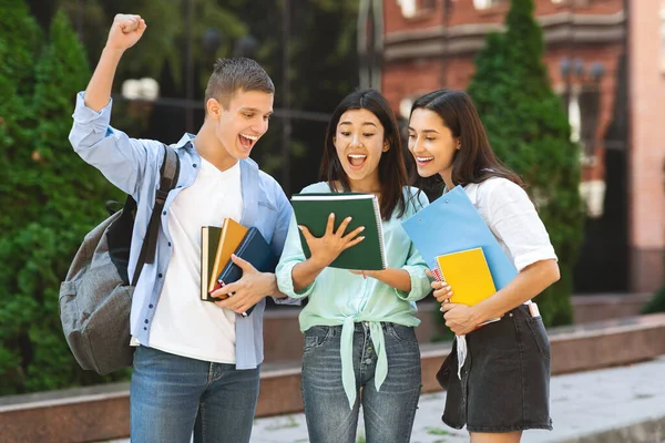 Allegri studenti universitari che leggono esami risultati in piedi nel campus, celebrando il successo — Foto Stock