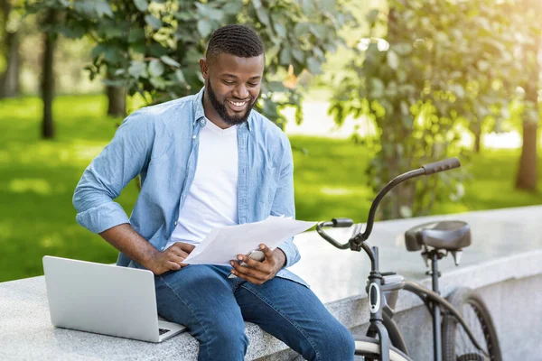 Homem preto feliz com laptop e bicicleta verificando documentação de negócios ao ar livre — Fotografia de Stock