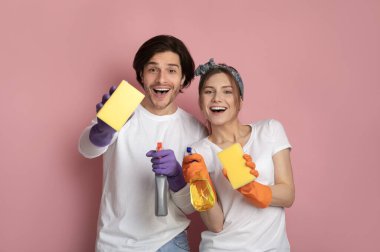 Pembe arka planda sprey deterjan ve temizlik süngeri olan mutlu bir çift.
