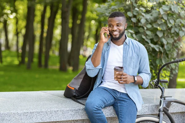 Bonito afro cara sentado no parque com café, falando no celular — Fotografia de Stock