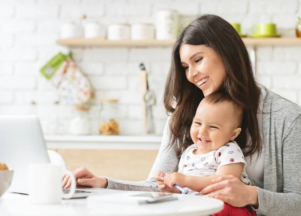 Happy woman working on laptop with baby at kitchen — Stock Photo, Image