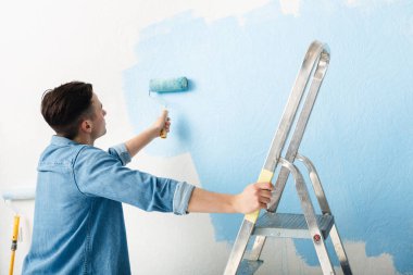 Young man leans on stepladder and paints wall