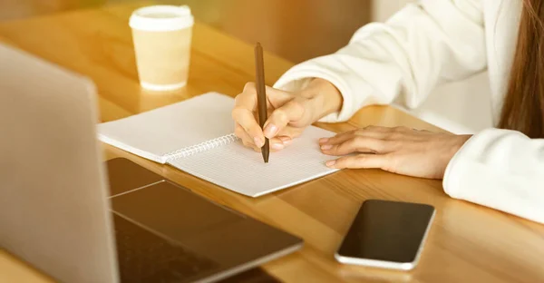 Girl writes in notebook on desk, near cup of coffee, phone and laptop — 스톡 사진