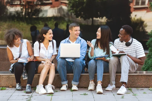 Estudiantes multiétnicos sentados al aire libre con computadora portátil, preparándose para clases juntos —  Fotos de Stock