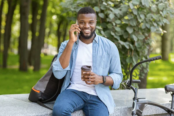 Hombre afroamericano bebiendo café en el parque y hablando por celular — Foto de Stock