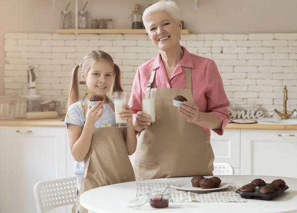 Cheerful grandmother and her lovely granddaughter with fresh muffins and milk in kitchen