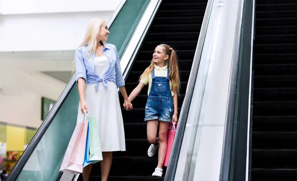 Mother And Daughter Moving Down Escalator Stairs In Mall