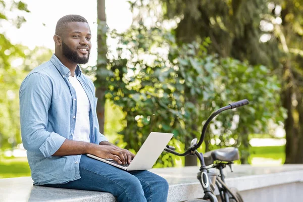 Pensivo homem africano sentado no parque com laptop, escrevendo ensaio ao ar livre — Fotografia de Stock