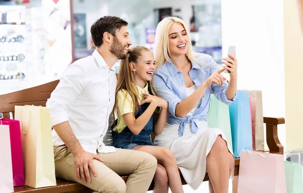Los padres y la hija usando el teléfono sentado en el banco en el hipermercado — Foto de Stock