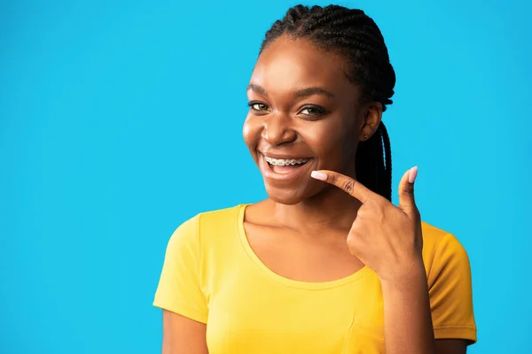 Africano senhora no aparelho apontando o dedo para os dentes, fundo azul — Fotografia de Stock