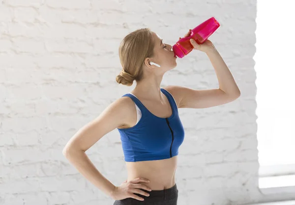 Chica joven en ropa deportiva beber agua de la botella contra la pared de ladrillo blanco — Foto de Stock
