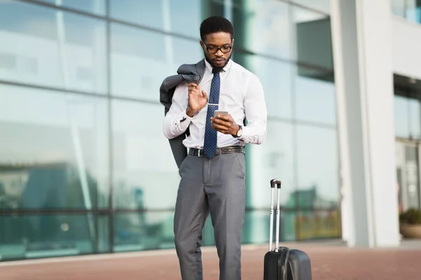 Imprenditore che utilizza il cellulare in piedi con valigia in aeroporto all'aperto — Foto Stock