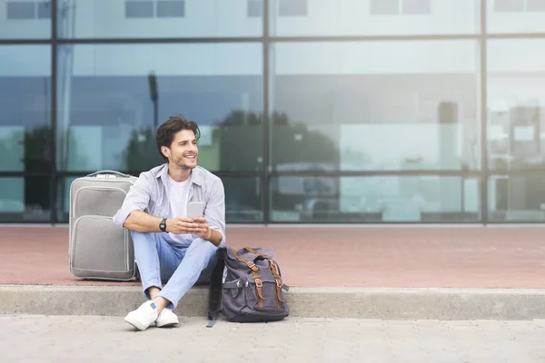 Hombre feliz sentado con equipaje y teléfono inteligente cerca de la terminal del aeropuerto — Foto de Stock