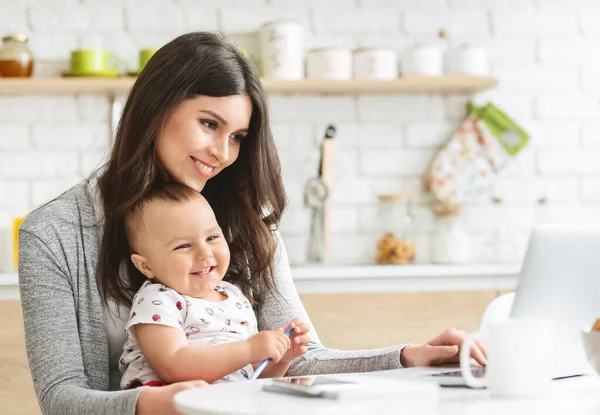 Jeune maman tapant sur ordinateur portable, assis avec bébé à la cuisine — Photo