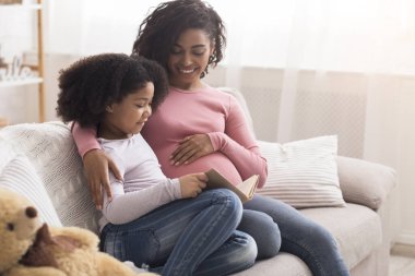 Adorable Little Girl Reading Book With Pregnant Mom In Living Room