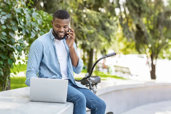 Empresário afro alegre falando no celular e usando laptop ao ar livre — Fotografia de Stock
