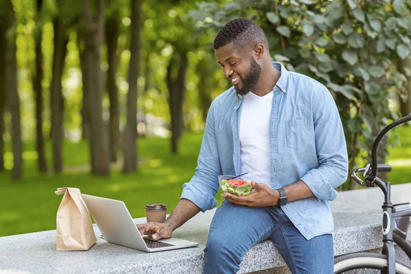 Joyful afro-americano cara usando laptop e almoçar ao ar livre — Fotografia de Stock