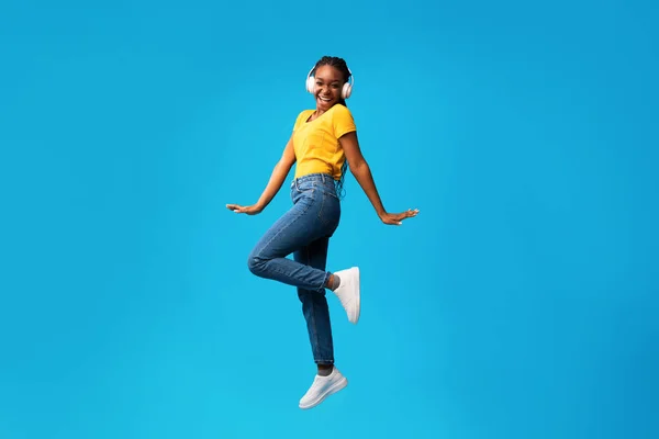 Mulher negra alegre em fones de ouvido pulando no fundo do estúdio azul — Fotografia de Stock