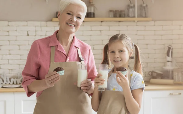 Ragazza carina con nonna che tiene muffin appena sfornati e bicchieri di latte in cucina — Foto Stock