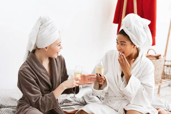 Twee jonge dames in badjassen zitten op bed champagne te drinken. — Stockfoto