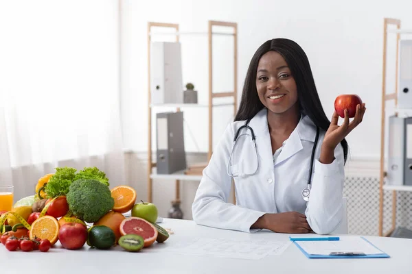 Medico afroamericano sorridente dietologo seduto alla scrivania, con mela in mano — Foto Stock