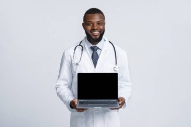 Beyaz cüppeli, siyah, neşeli doktor elinde boş laptop ekranı tutuyor.