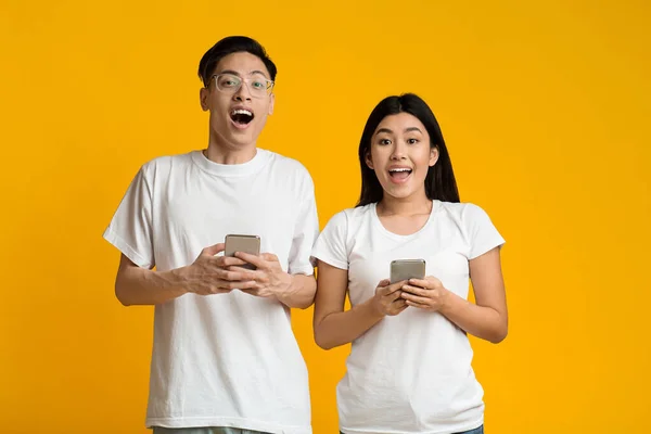 Heyecanlı genç Asyalı çift sarı arka planda akıllı telefonları tutuyor. — Stok fotoğraf