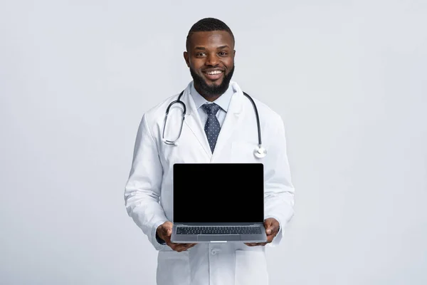 Beyaz cüppeli, siyah, neşeli doktor elinde boş laptop ekranı tutuyor. — Stok fotoğraf