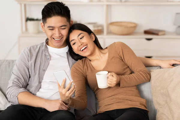 Asijský pár odpočívá na gauči, pomocí telefonu — Stock fotografie