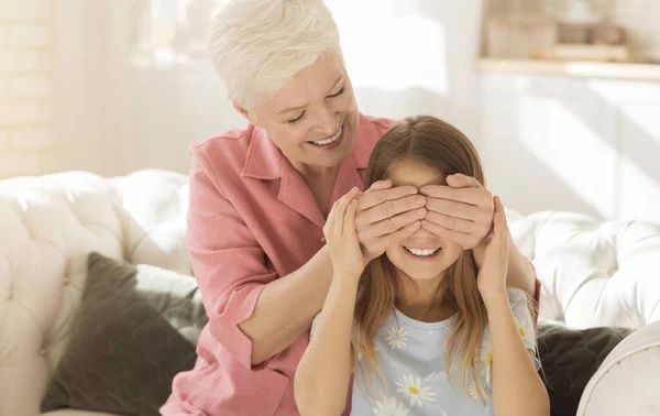 Playing Guess Who game. Cheerful elderly lady covering her granddaughters eyes, making surprise at home — Stock Photo, Image