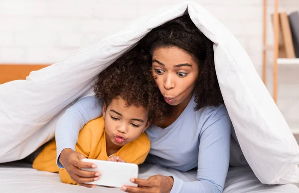 African Mom And Little Girl Playing Mobile Games In Bed