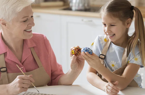 Family Easter traditions. Mature woman teaching her granddaughter how to paint eggs for spring holiday in kitchen — Stock Photo, Image