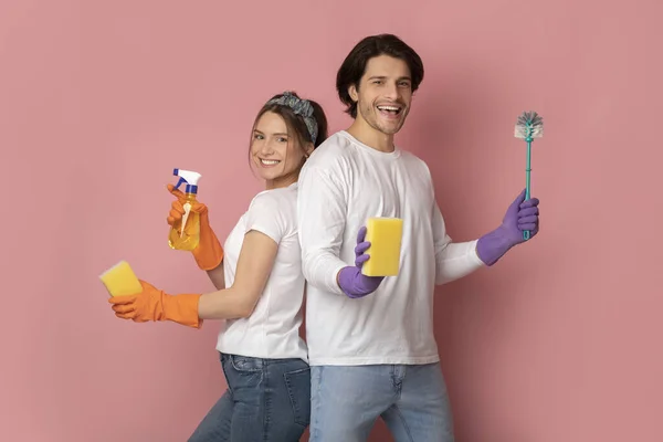 Joyful millennialcouple holding cleaning supplies posing over pink background in studio — Stock Photo, Image