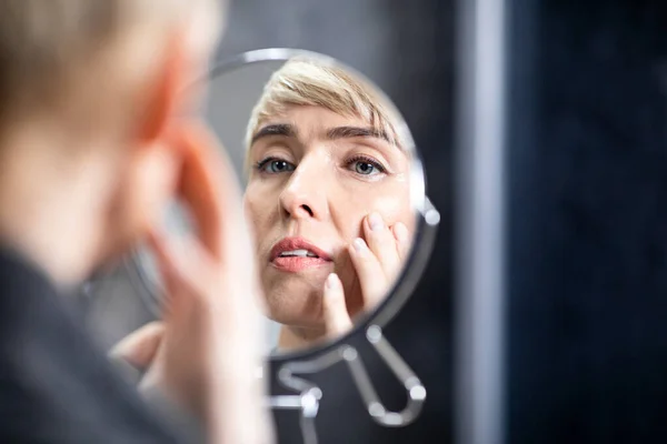 Dame mittleren Alters blickt im Spiegel auf Falten im Badezimmer drinnen — Stockfoto