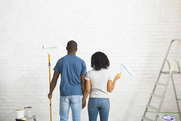 Αφροαμερικάνος άντρας και κορίτσι με ρολά στα χέρια βάφουν λευκό τοίχο από τούβλα — Φωτογραφία Αρχείου