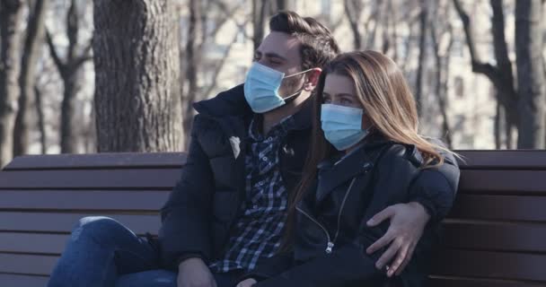 Jovem casal em máscaras protetoras sentado no banco no parque — Vídeo de Stock