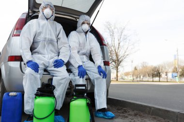 Biyolojik tehlike elbiseli adamlar dezenfeksiyon kimyasallarıyla arabada oturuyorlar.