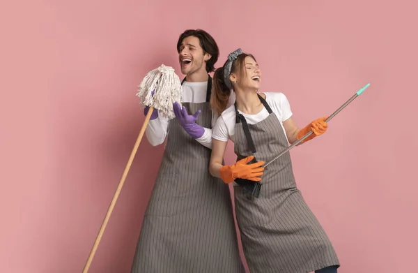 Pareja milenaria divirtiéndose durante la limpieza de primavera, usando escoba como guitarra de aire y cantando en fregona — Foto de Stock