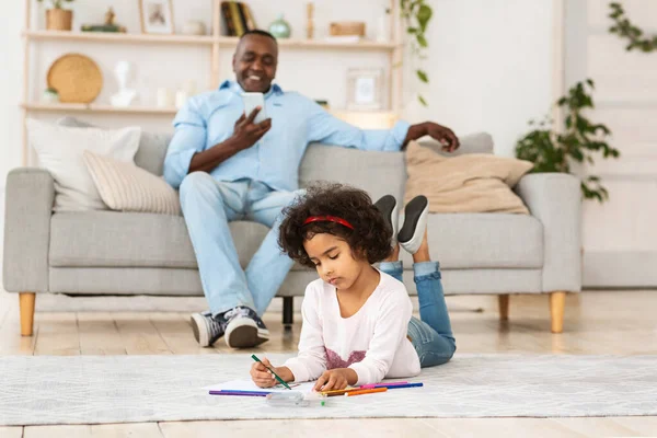Fique em casa. Menina afro-americana desenho no chão, enquanto seu avô usando smartphone dentro de casa — Fotografia de Stock