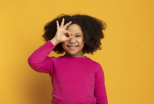 Carino poco afro ragazza mostrando ok gesto, guardando attraverso ok segno — Foto Stock