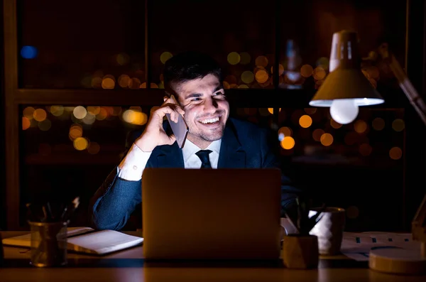 Gioioso uomo d'affari avendo conversazione telefonica seduto durante la notte sul posto di lavoro — Foto Stock