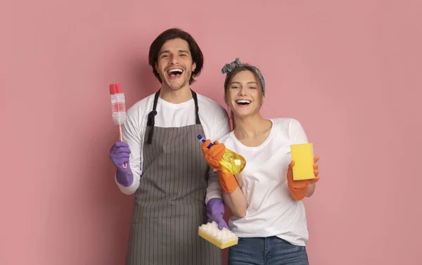 Веселая миллениальная пара с чистящими средствами в руках на розовом фоне — стоковое фото