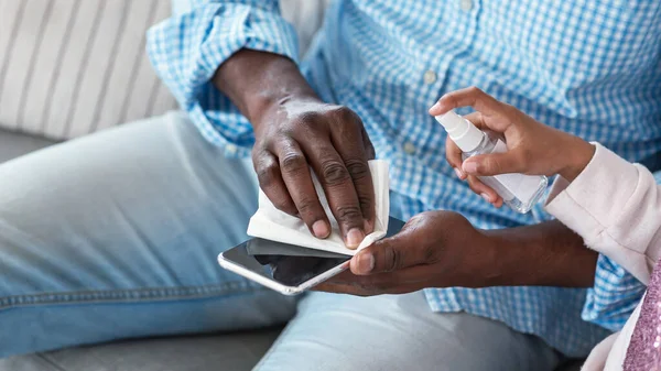 Enfeksiyon önleme. Afrika kökenli Amerikalı bir adam ve küçük bir çocuk cep telefonunu dezenfektanla temizliyor, yakın plan. Boş boşluk — Stok fotoğraf