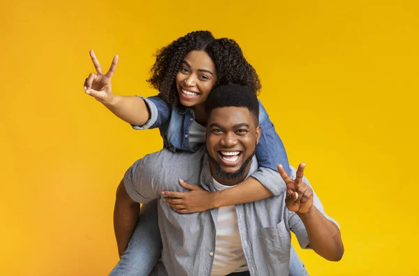 Joyful black couple embracing and showing peace sign, guy piggybacking girlfriend — Stock Photo, Image